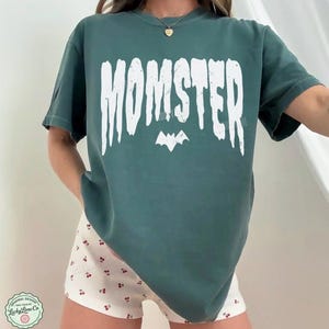May include: Blue spruce-colored t-shirt with the word "MOMSTER" in white, with a bat graphic underneath. The shirt is paired with white shorts patterned with red cherries. The shirt is from Lucky Lime Co.