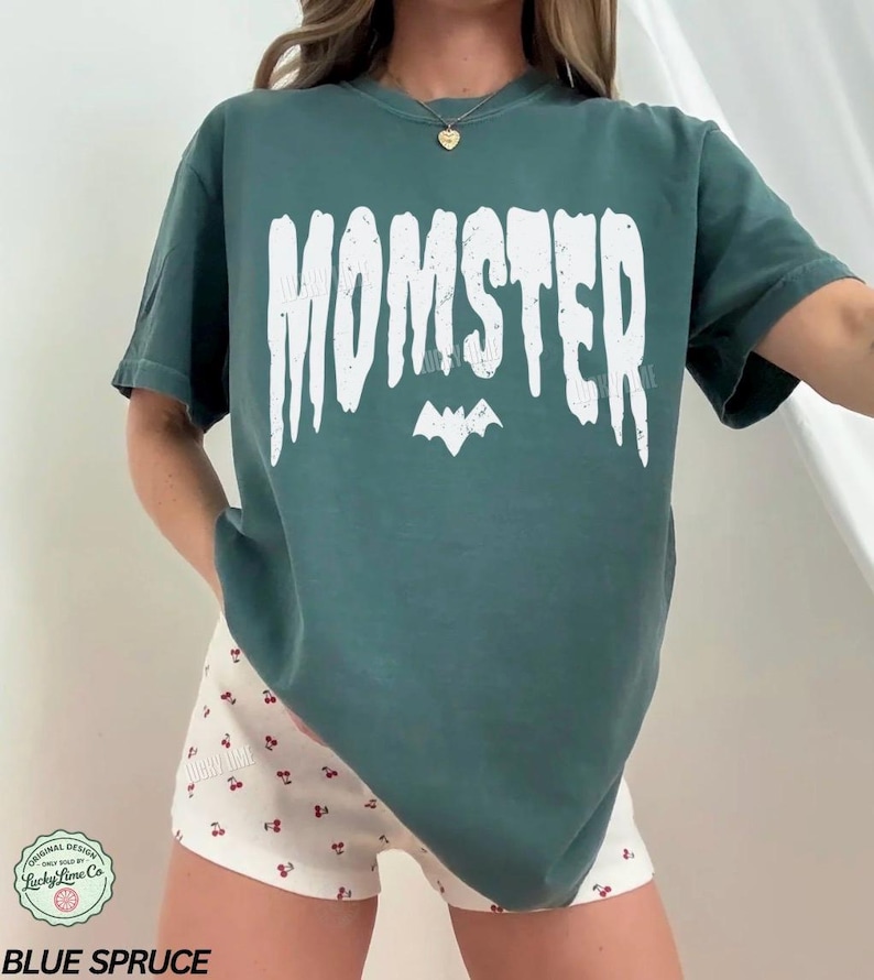 May include: Blue spruce-colored t-shirt with the word "MOMSTER" in white, with a bat graphic underneath. The shirt is paired with white shorts patterned with red cherries. The shirt is from Lucky Lime Co.