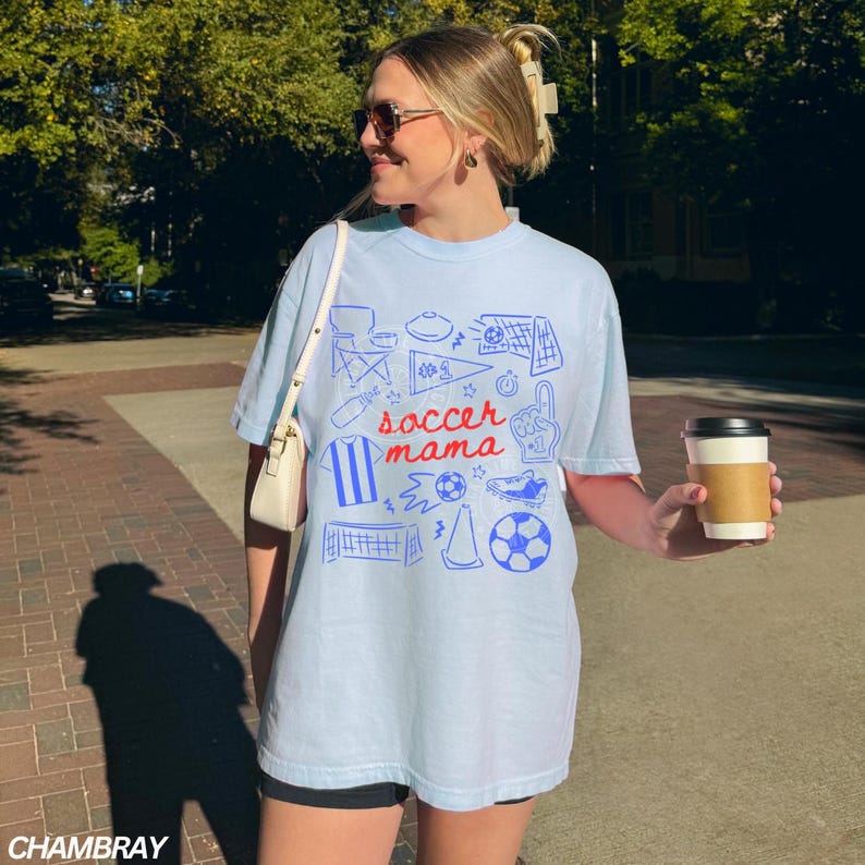 May include: Light blue t-shirt with a blue graphic design featuring soccer-related icons and the words "soccer mama." The shirt is a relaxed fit and has short sleeves. The person is holding a coffee cup.