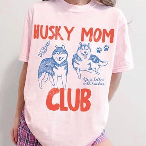 K&ouml;nnte beinhalten: Hellrosa T-Shirt mit dem Schriftzug "HUSKY MOM CLUB" in roten Buchstaben. Das Design zeigt blaue Illustrationen von zwei Huskys, Pfotenabdr&uuml;cken und den Satz "life is better with huskies".