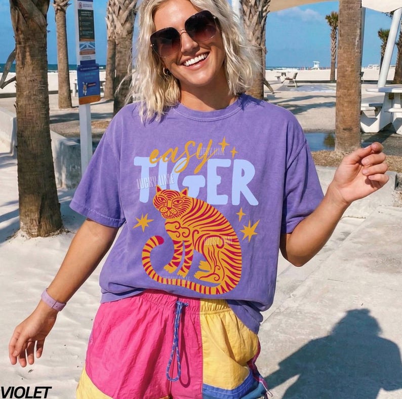 May include: A purple t-shirt with the text "easy + TIGER" and a tiger illustration in orange and yellow. The person is wearing pink, yellow, and blue shorts. The background includes palm trees and a beach.