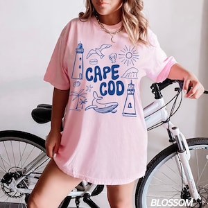Peut inclure: Un t-shirt rose avec un motif graphique bleu qui dit "Cape Cod". Le graphique comprend un phare, une baleine, une coquille, une &eacute;toile de mer, un palmier, un soleil et une vague.