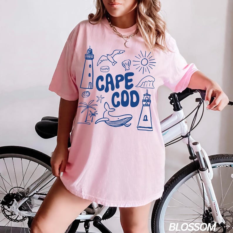 Peut inclure: Un t-shirt rose avec un motif graphique bleu qui dit "Cape Cod". Le graphique comprend un phare, une baleine, une coquille, une &eacute;toile de mer, un palmier, un soleil et une vague.