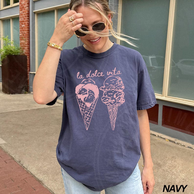 Op de afbeelding: Een marineblauw t-shirt met een roze afbeelding van twee ijshoorns. De tekst "la dolce vita" is boven de ijshoorns gedrukt.