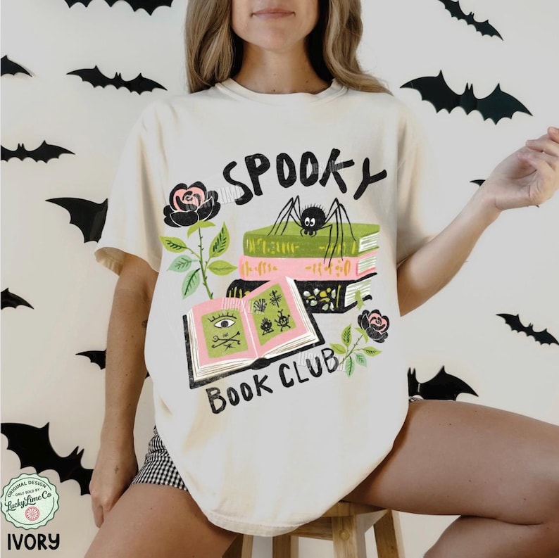 Op de afbeelding: Ivoor kleurig T-shirt met een "Spooky Book Club" ontwerp. De afbeelding toont een spin op boeken, open boeken met oog- en plant illustraties en rozen. Het shirt heeft korte mouwen en een relaxte pasvorm.