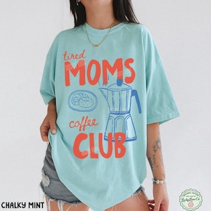 May include: A light mint green t-shirt featuring a graphic print in red and blue. The design includes the text "tired MOMS coffee CLUB" alongside an illustration of a moka pot and a small plate with a pastry. The shirt has short sleeves.