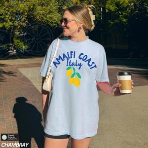 K&ouml;nnte beinhalten: Ein hellblaues T-Shirt mit dem Text "Amalfi Coast Italy" in blauer Schrift und einer Grafik von Zitronen. Das Shirt ist Oversize und hat eine lockere Passform.