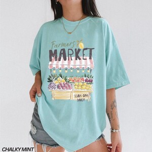 May include: A light blue t-shirt with a graphic of a farmer's market stand. The stand is filled with colorful fruits and vegetables. The text "Farmers Market" is above the stand, and "10AM-5PM DAILY" is below.