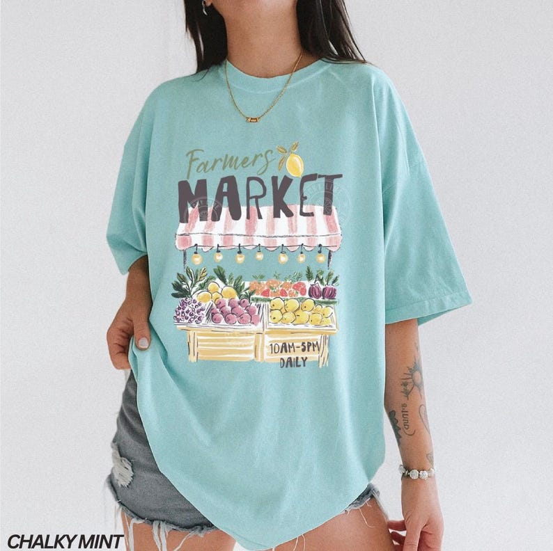 May include: A light blue t-shirt with a graphic of a farmer's market stand. The stand is filled with colorful fruits and vegetables. The text "Farmers Market" is above the stand, and "10AM-5PM DAILY" is below.