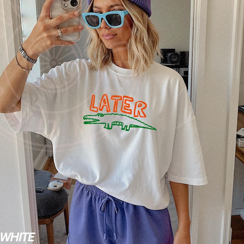 May include: White oversized t-shirt with a green and orange alligator graphic and the word "LATER" printed on the front.