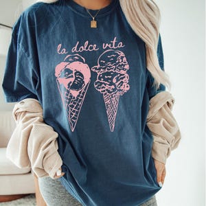 Op de afbeelding: Een marineblauw t-shirt met een roze afbeelding van twee ijshoorns en de tekst "la dolce vita".