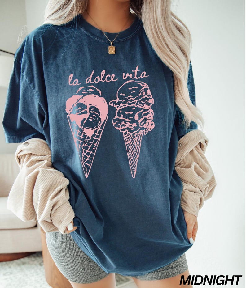 Op de afbeelding: Een marineblauw t-shirt met een roze afbeelding van twee ijshoorns en de tekst "la dolce vita".