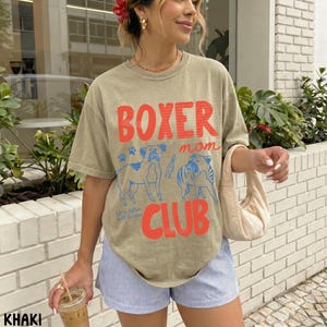 May include: A khaki short-sleeve t-shirt featuring the red text BOXER CLUB and a blue graphic of two boxer dogs. The shirt includes the text mom in red script and the phrase life is better with boxers in small blue text. The shirt is worn with blue and white striped shorts.