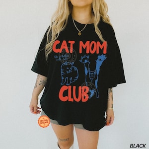 Puede incluir: Camiseta negra de gran tama&ntilde;o con las palabras "CAT MOM CLUB" en rojo y azul. La camiseta presenta ilustraciones de gatos y la frase "life is better with cats". Es visible una pegatina redonda de "Original Designer".