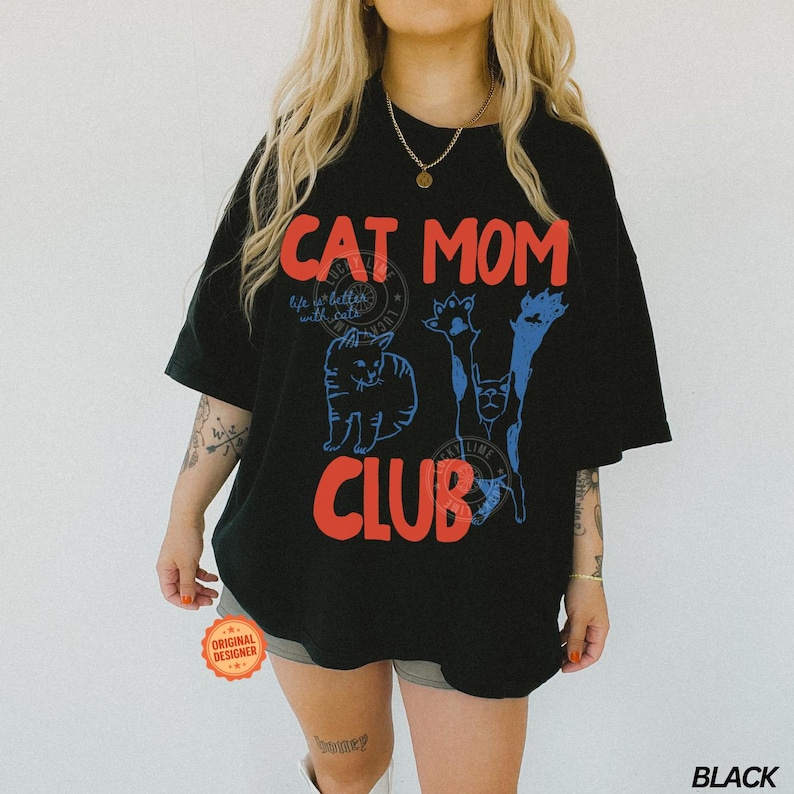 Puede incluir: Camiseta negra de gran tama&ntilde;o con las palabras "CAT MOM CLUB" en rojo y azul. La camiseta presenta ilustraciones de gatos y la frase "life is better with cats". Es visible una pegatina redonda de "Original Designer".
