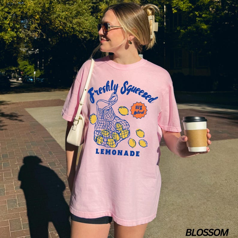 Puede incluir: Camiseta rosa con un gr&aacute;fico de una bolsa de malla llena de limones y el texto "Freshly Squeezed 100% Natural LEMONADE".
