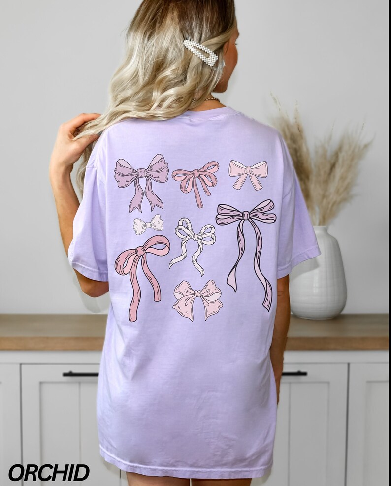 May include: A lavender t-shirt with a graphic of pink and white bows on the back. The shirt is a relaxed fit and has a crew neck.