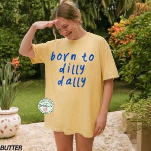 May include: Butter yellow t-shirt with the phrase "born to dilly dally" in blue script. The shirt has a relaxed fit and a crew neck. The design includes a circular logo from Lucky Lime Co.