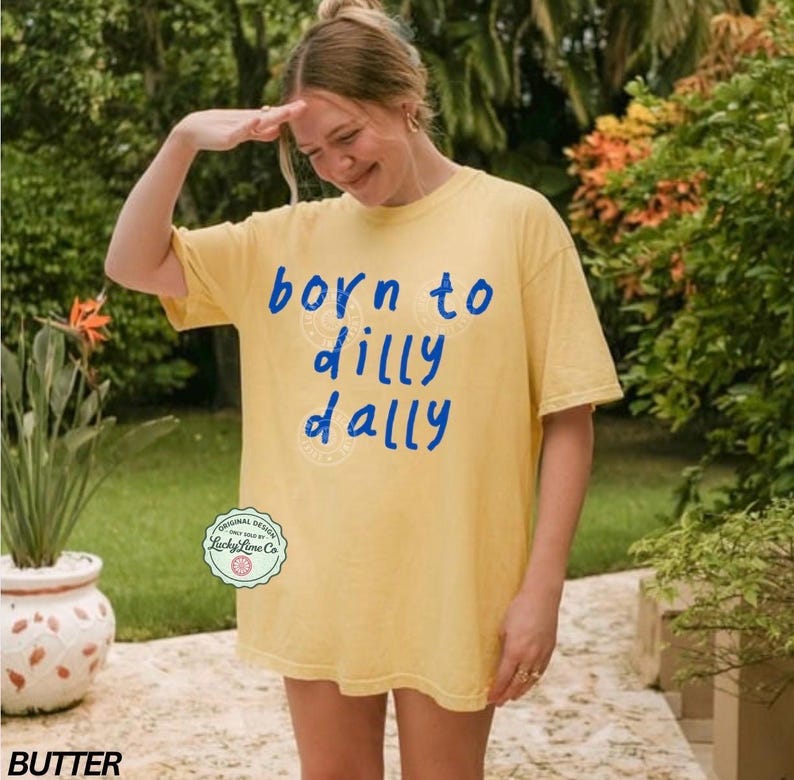 May include: Butter yellow t-shirt with the phrase "born to dilly dally" in blue script. The shirt has a relaxed fit and a crew neck. The design includes a circular logo from Lucky Lime Co.
