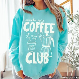 May include: A turquoise sweatshirt with the words "pumpkin spice COFFEE all every day day CLUB" in white. The design includes a coffee cup and a coffee maker illustration. The sweatshirt is a comfortable, casual style.
