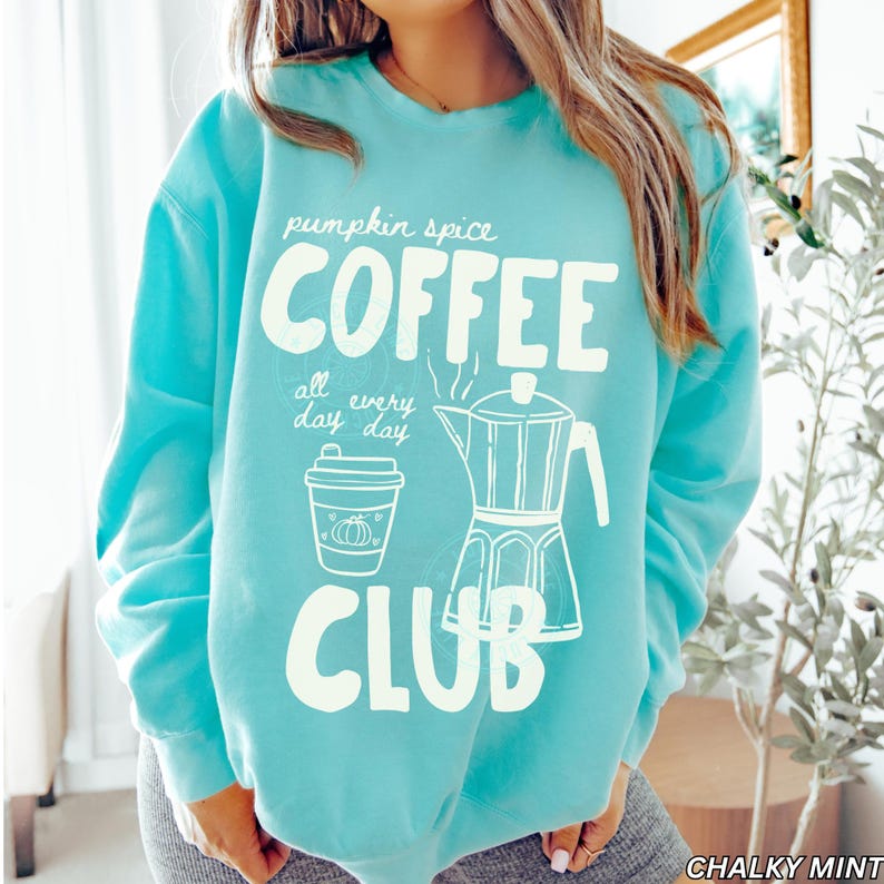 May include: A turquoise sweatshirt with the words "pumpkin spice COFFEE all every day day CLUB" in white. The design includes a coffee cup and a coffee maker illustration. The sweatshirt is a comfortable, casual style.