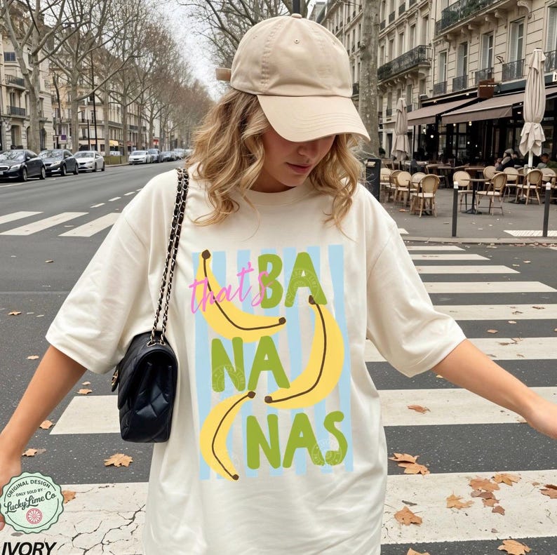 May include: Ivory-colored t-shirt with the text "That's Bananas" in pink, yellow, and green. The design includes yellow banana illustrations on a light blue striped background. The shirt is worn with a beige baseball cap.
