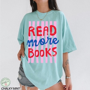 Puede incluir: Camiseta azul claro con las palabras "READ more BOOKS" en rojo y azul, sobre un fondo de rayas rosas y blancas. La camiseta tiene un corte holgado y mangas cortas. La marca "Chalky Mint" est&aacute; en la parte inferior.