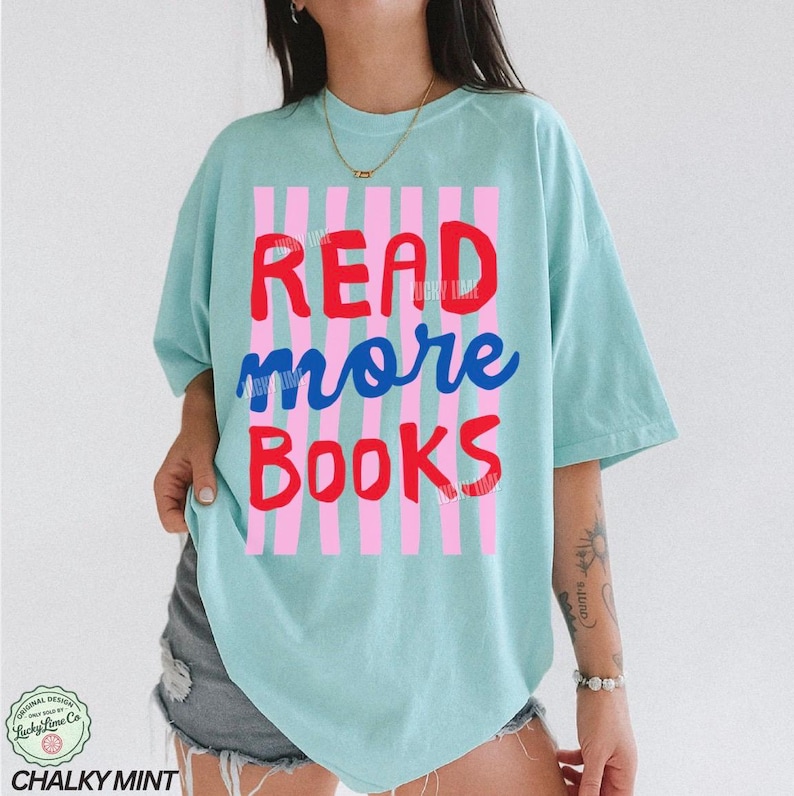 Puede incluir: Camiseta azul claro con las palabras "READ more BOOKS" en rojo y azul, sobre un fondo de rayas rosas y blancas. La camiseta tiene un corte holgado y mangas cortas. La marca "Chalky Mint" est&aacute; en la parte inferior.
