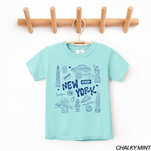 May include: A light turquoise t-shirt with a blue graphic of New York City landmarks and text. The design includes the Brooklyn Bridge, the Statue of Liberty, and the Empire State Building. The shirt is hanging on a wooden rack.
