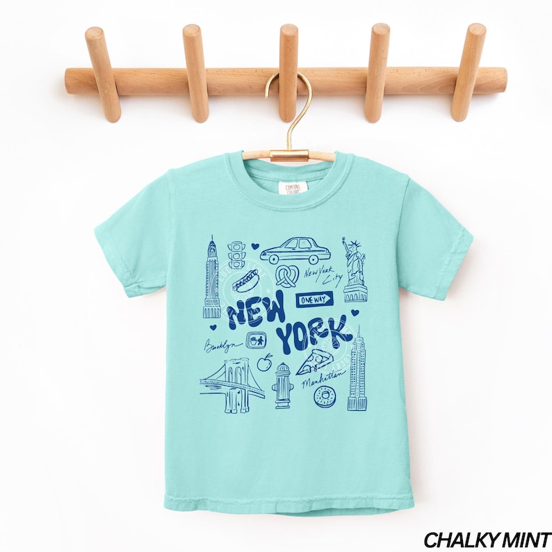 May include: A light turquoise t-shirt with a blue graphic of New York City landmarks and text. The design includes the Brooklyn Bridge, the Statue of Liberty, and the Empire State Building. The shirt is hanging on a wooden rack.