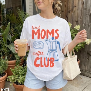May include: A white short-sleeve t-shirt featuring the red text "tired MOMS" above a blue line drawing of a plate with a pastry and a moka pot, with the blue text "coffee" and red text "CLUB" below, paired with light blue pinstriped shorts.
