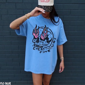 May include: A light blue t-shirt featuring a black graphic of a skull with pink flames emerging from the eye sockets. Below the skull, the text reads "EVERYTHIN'G'S FINE" in black lettering. The shirt has short sleeves and a crew neckline.