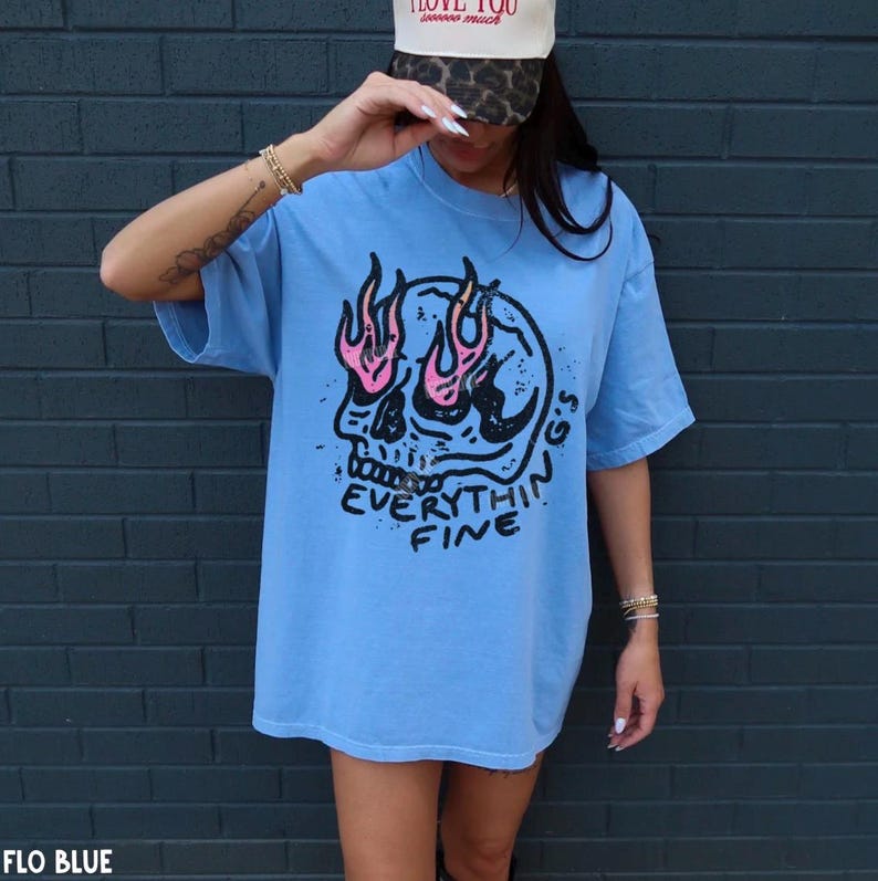 May include: A light blue t-shirt featuring a black graphic of a skull with pink flames emerging from the eye sockets. Below the skull, the text reads "EVERYTHIN'G'S FINE" in black lettering. The shirt has short sleeves and a crew neckline.
