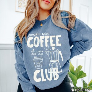 May include: Blue jean colored sweatshirt with the words "pumpkin spice COFFEE CLUB" in white block letters. The design includes a coffee cup with a pumpkin and a coffee pot. The text "all every day day" is also included.