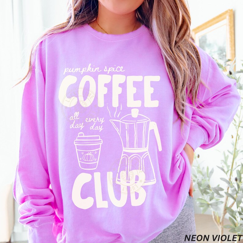 May include: A neon violet sweatshirt with the words "pumpkin spice COFFEE CLUB" in white. The design includes a coffee cup, a coffee maker, and the phrase "all every day". The sweatshirt is a comfortable, casual style.