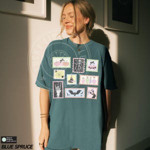 May include: Blue Spruce t-shirt featuring a collection of Halloween-themed postage stamp designs. The stamps include images of pumpkins, skeletons, bats, and potions. The shirt has short sleeves and a relaxed fit. The text "BLUE SPRUCE" is at the bottom.