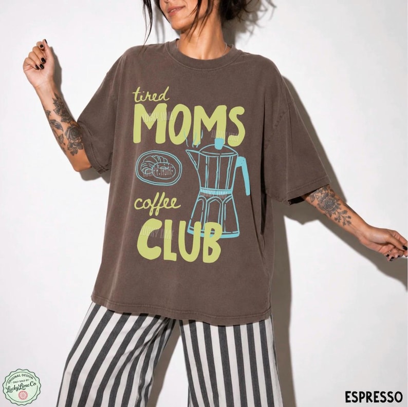 May include: A dark brown short-sleeve t-shirt featuring the text "tired MOMS coffee CLUB" in light green. The graphic includes a light blue outline of a moka pot and a small pastry on a plate. The shirt has a crew neckline and a loose fit.