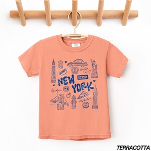 May include: Terracotta-colored t-shirt with a blue graphic of New York City landmarks, including the Statue of Liberty, the Empire State Building, and the Brooklyn Bridge. The words "New York" are prominently displayed.