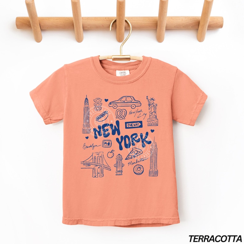 May include: Terracotta-colored t-shirt with a blue graphic of New York City landmarks, including the Statue of Liberty, the Empire State Building, and the Brooklyn Bridge. The words "New York" are prominently displayed.