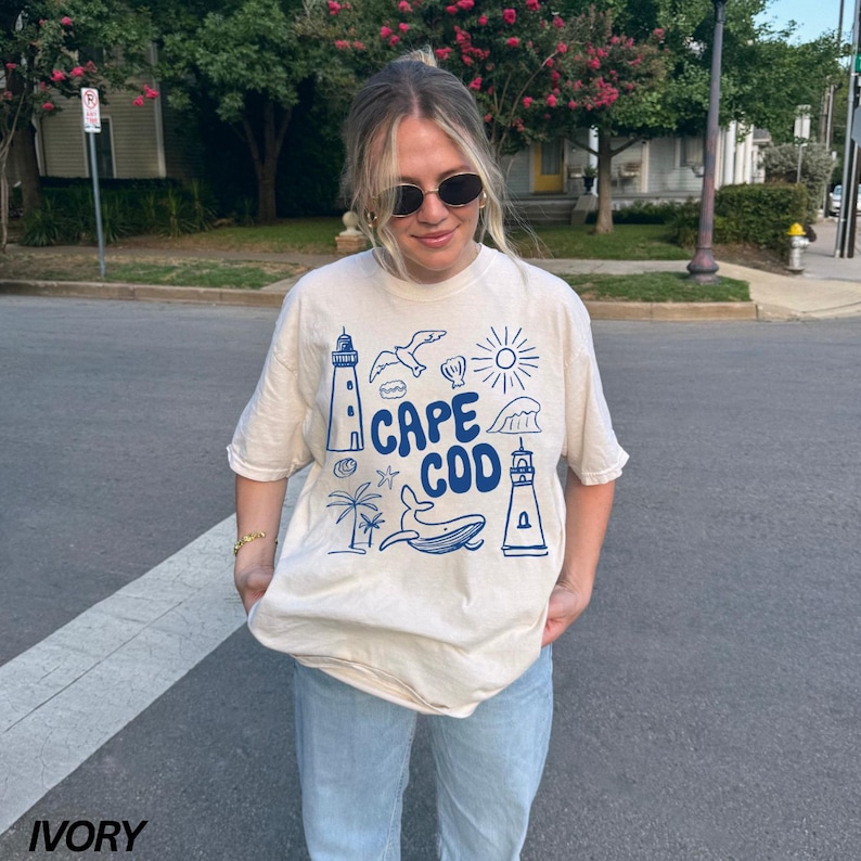 Peut inclure: Un t-shirt beige clair avec un motif graphique bleu repr&eacute;sentant un phare, une baleine, le soleil et des vagues. Le texte "Cape Cod" est &eacute;crit en bleu sur le t-shirt.