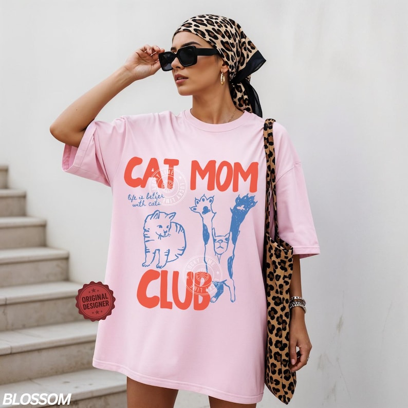Puede incluir: Una camiseta rosa claro de gran tama&ntilde;o con las palabras "CAT MOM CLUB" en rojo y azul, junto con ilustraciones de gatos. La camiseta tambi&eacute;n presenta el texto "life is better with cats". Se ve un bolso tote con estampado de leopardo.