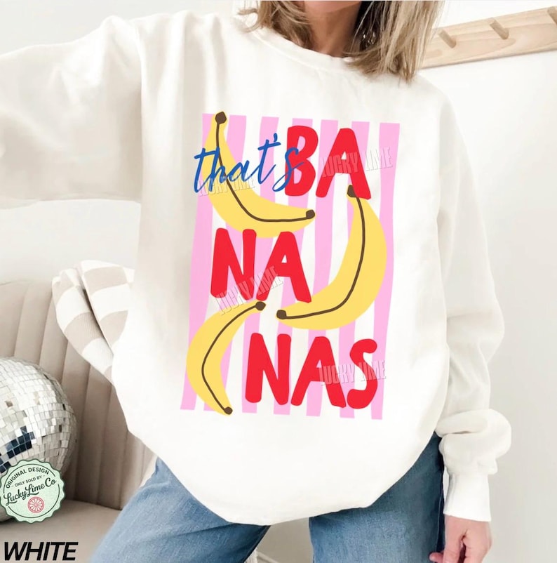 May include: White sweatshirt with the phrase "that's BANANAS" in red and blue text, with yellow banana illustrations on a pink and white striped background. The sweatshirt is a long-sleeved crew neck.