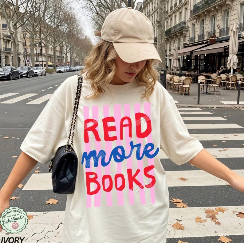 Puede incluir: Camiseta de color marfil con el texto "READ more BOOKS" en rojo y azul, sobre rayas verticales rosas. La persona lleva una gorra de b&eacute;isbol beige y un bolso negro con correa de cadena.