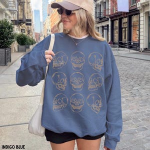 May include: An indigo blue crewneck sweatshirt featuring a repeating grid pattern of nine gold-colored line art skulls. The long-sleeved garment has a relaxed fit and a ribbed neckline, cuffs, and hem, showcasing a graphic print of human skulls.