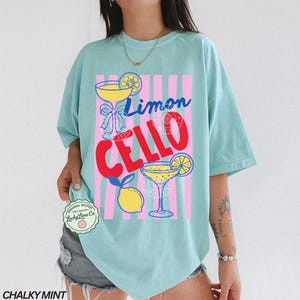 May include: A light blue t-shirt with a graphic design featuring the words "Limon Cello" in blue and red, along with illustrations of cocktails and lemons. The shirt has a relaxed fit.