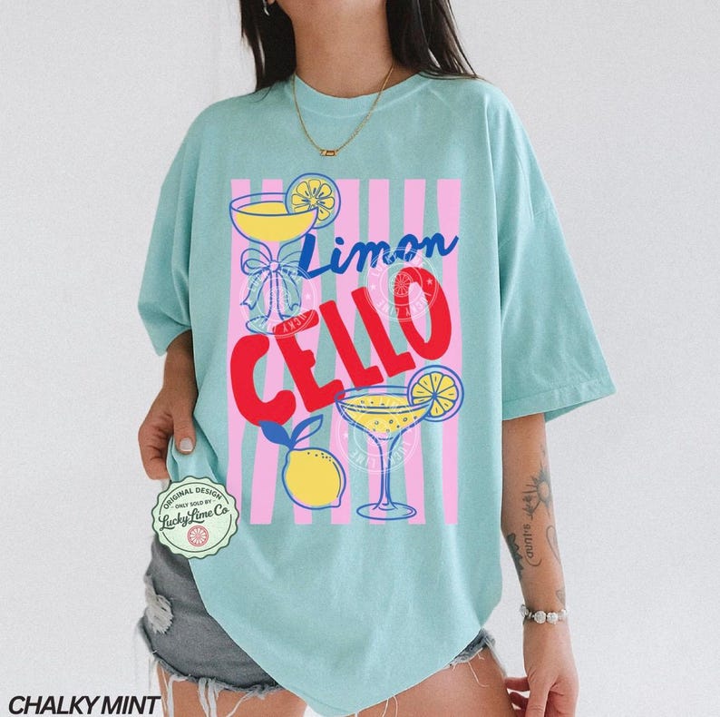 May include: A light blue t-shirt with a graphic design featuring the words "Limon Cello" in blue and red, along with illustrations of cocktails and lemons. The shirt has a relaxed fit.