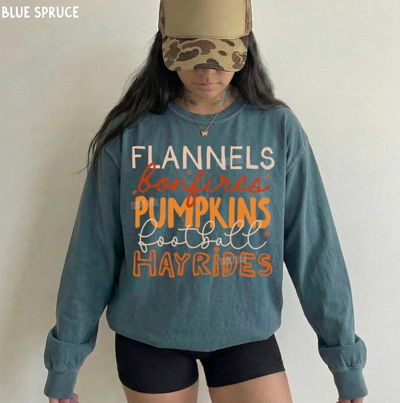 May include: A blue-green long-sleeve sweatshirt featuring the text FLANNELS, bonfires, PUMPKINS, football, and HAYRIDES in white and orange lettering. The garment is paired with a camouflage baseball cap with a tan front panel and a gold necklace.