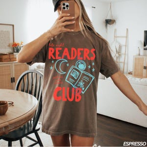 May include: Espresso-colored t-shirt with a graphic design. The design features the words "late night READERS CLUB" in red, with a blue crescent moon, stars, and an open book illustration. The shirt is a casual, oversized fit.