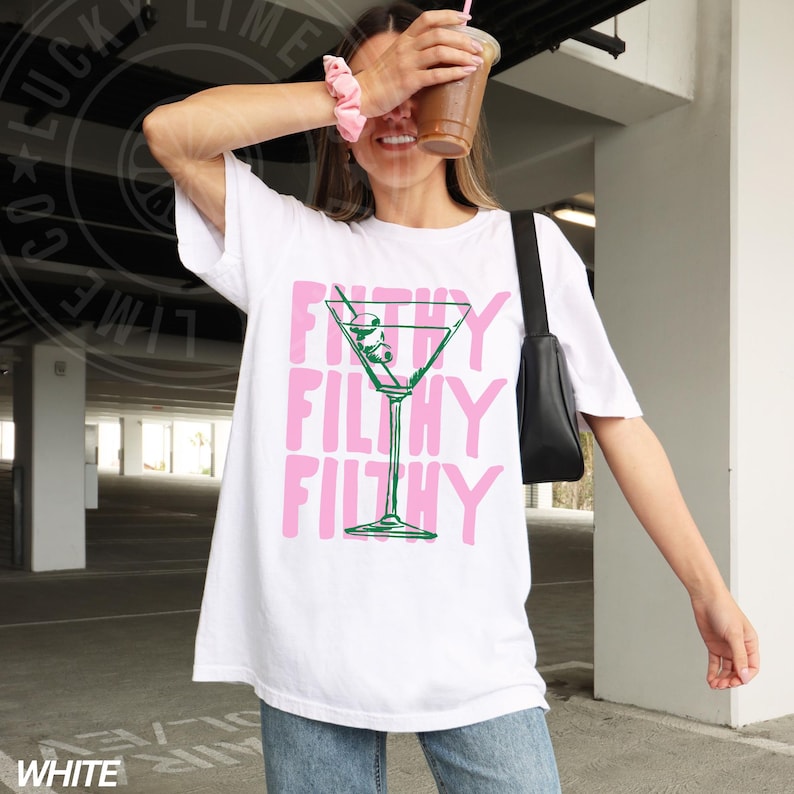 Pode incluir: Camiseta branca oversized com um gr&aacute;fico rosa de um copo de martini com azeitonas e o texto "Filthy Filthy Filthy".