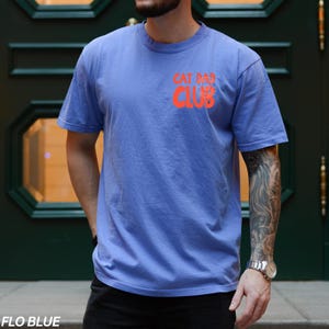 Peut inclure: T-shirt bleu avec le texte "CAT DAD CLUB" en orange sur la poitrine gauche. Le t-shirt est de couleur bleu uni et a des manches courtes. Le texte "FLO BLUE" est en bas.